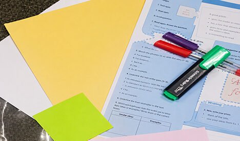 A worksheet, coloured paper and sticky notes arranged on a table, alongside several coloured highlighter pens, ready for a classroom or study activity.