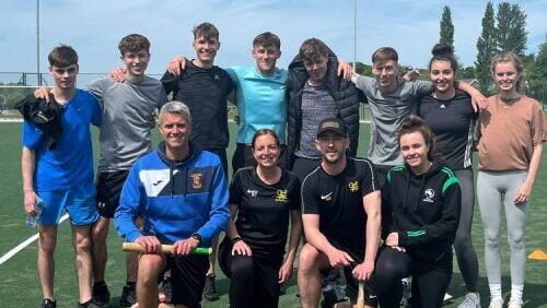 A group of students and staff posing for a photograph on an outdoor sports pitch, with some standing and others seated at the front, holding hockey sticks and wearing sports clothing, with trees and sports fencing visible in the background.