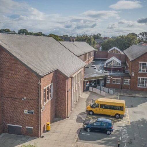 Aerial view of the Castell Alun school campus, showing red brick school buildings, a car park with vehicles including a yellow minibus, and surrounding trees beneath a partly cloudy sky.