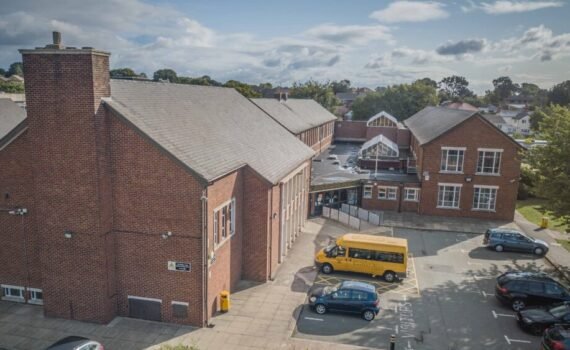 Aerial view of the Castell Alun school campus, showing red brick school buildings, a car park with vehicles including a yellow minibus, and surrounding trees beneath a partly cloudy sky.