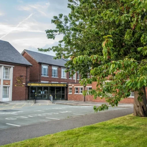 Front entrance of the Castell Alun school building, with red brick and white-panelled façades, a large tree and grassy area in the foreground, and the school name displayed above the main entrance.