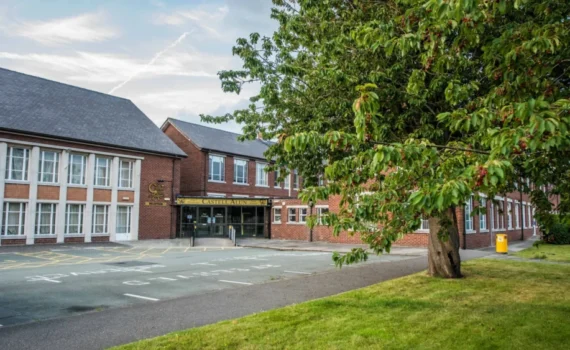 Front entrance of the Castell Alun school building, with red brick and white-panelled façades, a large tree and grassy area in the foreground, and the school name displayed above the main entrance.