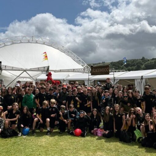 A large group of students in black school sportswear gathered on a grassy field at an outdoor event, posing for a group photograph between white marquee tents, with a large arched structure and cloudy sky in the background.