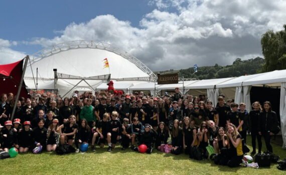 A large group of students in black school sportswear gathered on a grassy field at an outdoor event, posing for a group photograph between white marquee tents, with a large arched structure and cloudy sky in the background.