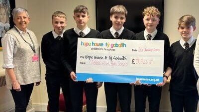 Five students in school uniform standing alongside an adult, holding a large Hope House and Tŷ Gobaith Children's Hospices charity cheque, posed indoors in front of a display screen.