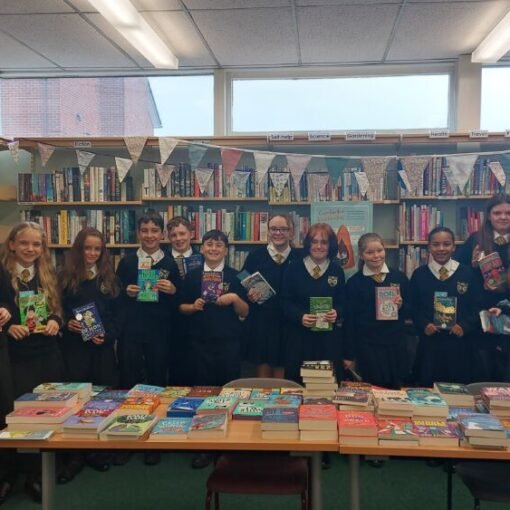 A group of students in school uniform standing in a library behind tables displaying a wide selection of books, each holding a book, with bookshelves and bunting visible in the background.