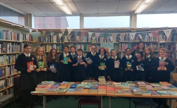 A group of students in school uniform standing in a library behind tables displaying a wide selection of books, each holding a book, with bookshelves and bunting visible in the background.