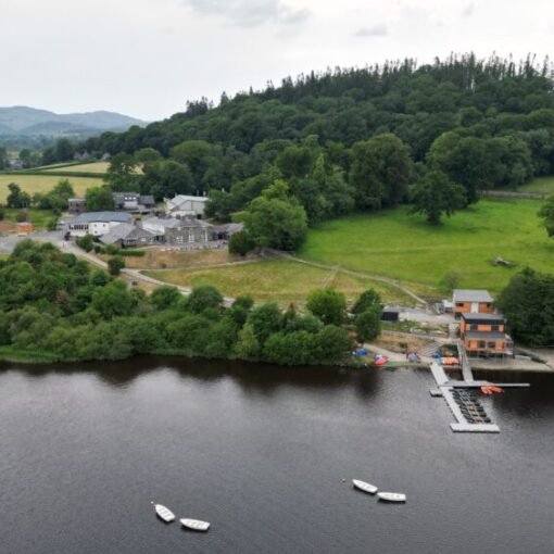 Aerial view of a lakeside outdoor activity centre surrounded by woodland and open countryside, featuring several buildings, a jetty extending into the water, small boats moored on the lake, and rolling hills in the background.