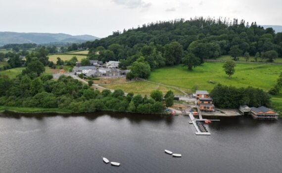 Aerial view of a lakeside outdoor activity centre surrounded by woodland and open countryside, featuring several buildings, a jetty extending into the water, small boats moored on the lake, and rolling hills in the background.