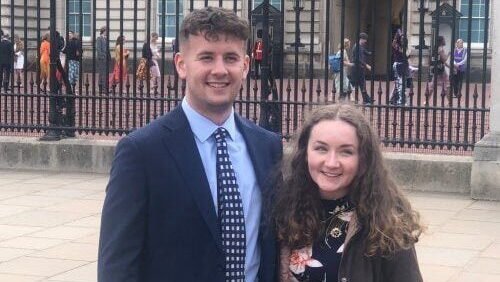 Two people standing outdoors in front of a historic building and metal railings, posing for a photograph, with pedestrians visible in the background.