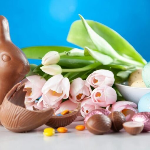 Chocolate Easter bunny surrounded by pastel-coloured decorated eggs, pink tulips and pieces of broken chocolate on a table, set against a bright blue background.