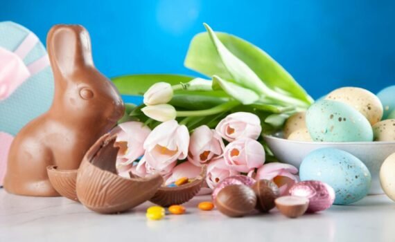 Chocolate Easter bunny surrounded by pastel-coloured decorated eggs, pink tulips and pieces of broken chocolate on a table, set against a bright blue background.