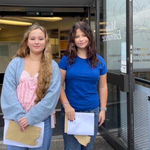 Two students standing outside the entrance to Castell Alun High School, holding envelopes and papers while posing for a photograph in front of the school's glass entrance doors and signage.