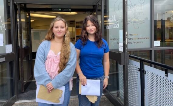 Two students standing outside the entrance to Castell Alun High School, holding envelopes and papers while posing for a photograph in front of the school's glass entrance doors and signage.