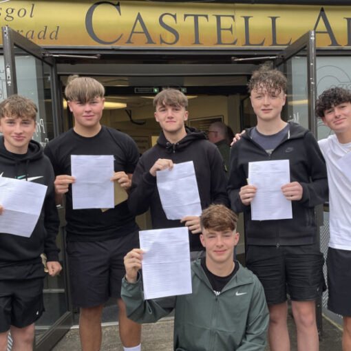 Six students standing outside the entrance to Castell Alun High School, holding sheets of paper and posing for a photograph beneath the school sign and open entrance doors.