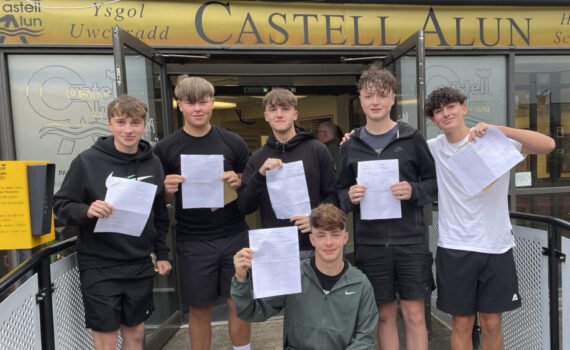 Six students standing outside the entrance to Castell Alun High School, holding sheets of paper and posing for a photograph beneath the school sign and open entrance doors.