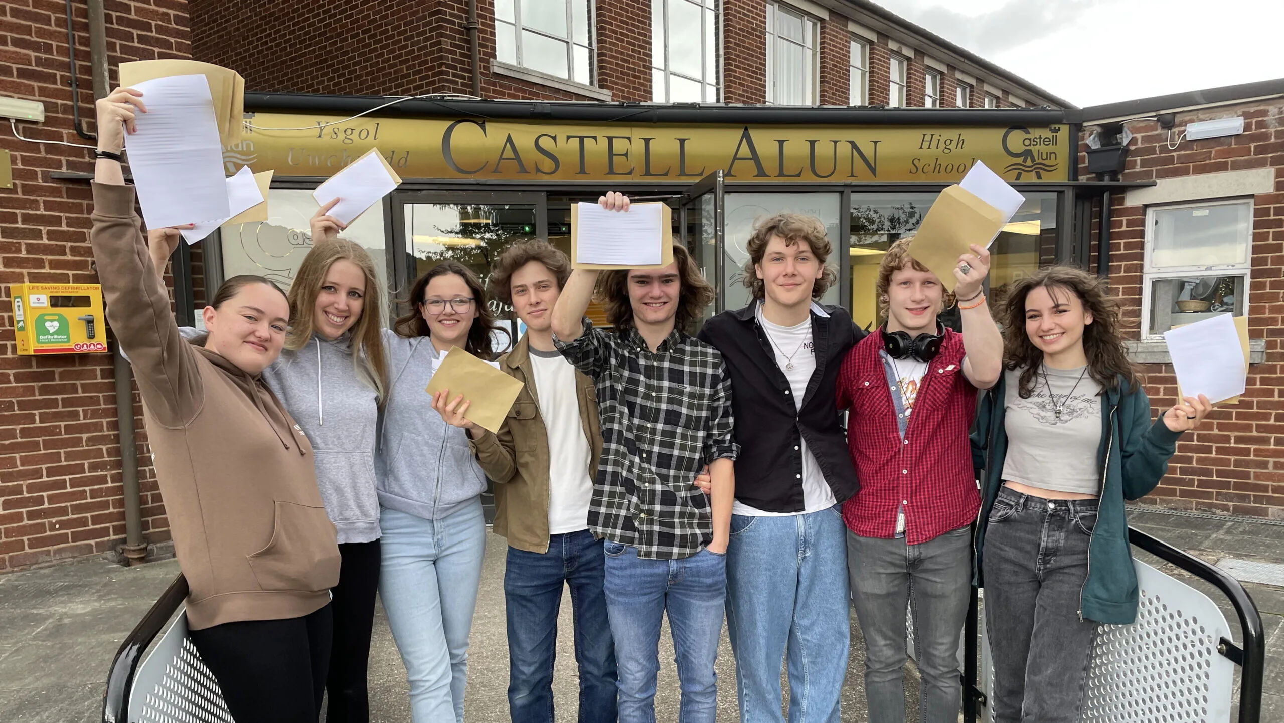 A group of students standing outside the main entrance of Castell Alun High School, holding envelopes and papers aloft while posing for a photograph beneath the school sign.