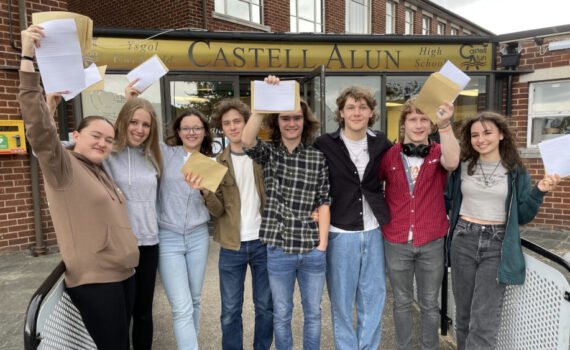 A group of students standing outside the main entrance of Castell Alun High School, holding envelopes and papers aloft while posing for a photograph beneath the school sign.