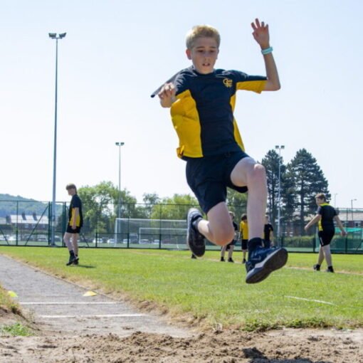 A student in a black and yellow sports kit competing in the long jump, airborne above the sand pit during an athletics event, with other students, sports fencing and floodlights visible in the background on a sunny day.