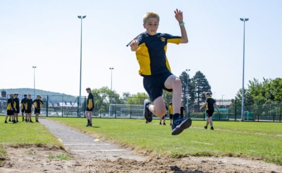 A student in a black and yellow sports kit competing in the long jump, airborne above the sand pit during an athletics event, with other students, sports fencing and floodlights visible in the background on a sunny day.