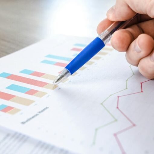 A hand holding a blue pen and pointing to printed business charts and graphs on a report, including coloured bar charts and line graphs displayed on a desk.