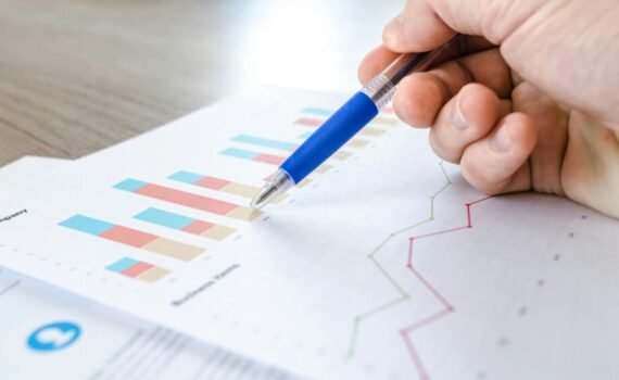 A hand holding a blue pen and pointing to printed business charts and graphs on a report, including coloured bar charts and line graphs displayed on a desk.