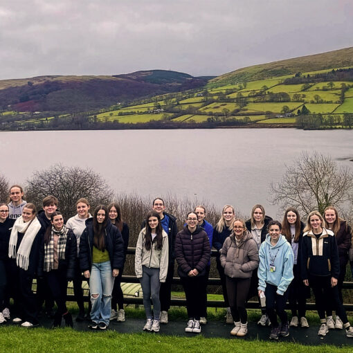 A large group of students and staff standing together by a lakeside viewpoint, with calm water in the foreground and rolling green hills and farmland in the background under an overcast sky.
