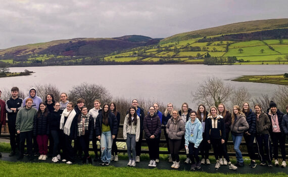 A large group of students and staff standing together by a lakeside viewpoint, with calm water in the foreground and rolling green hills and farmland in the background under an overcast sky.