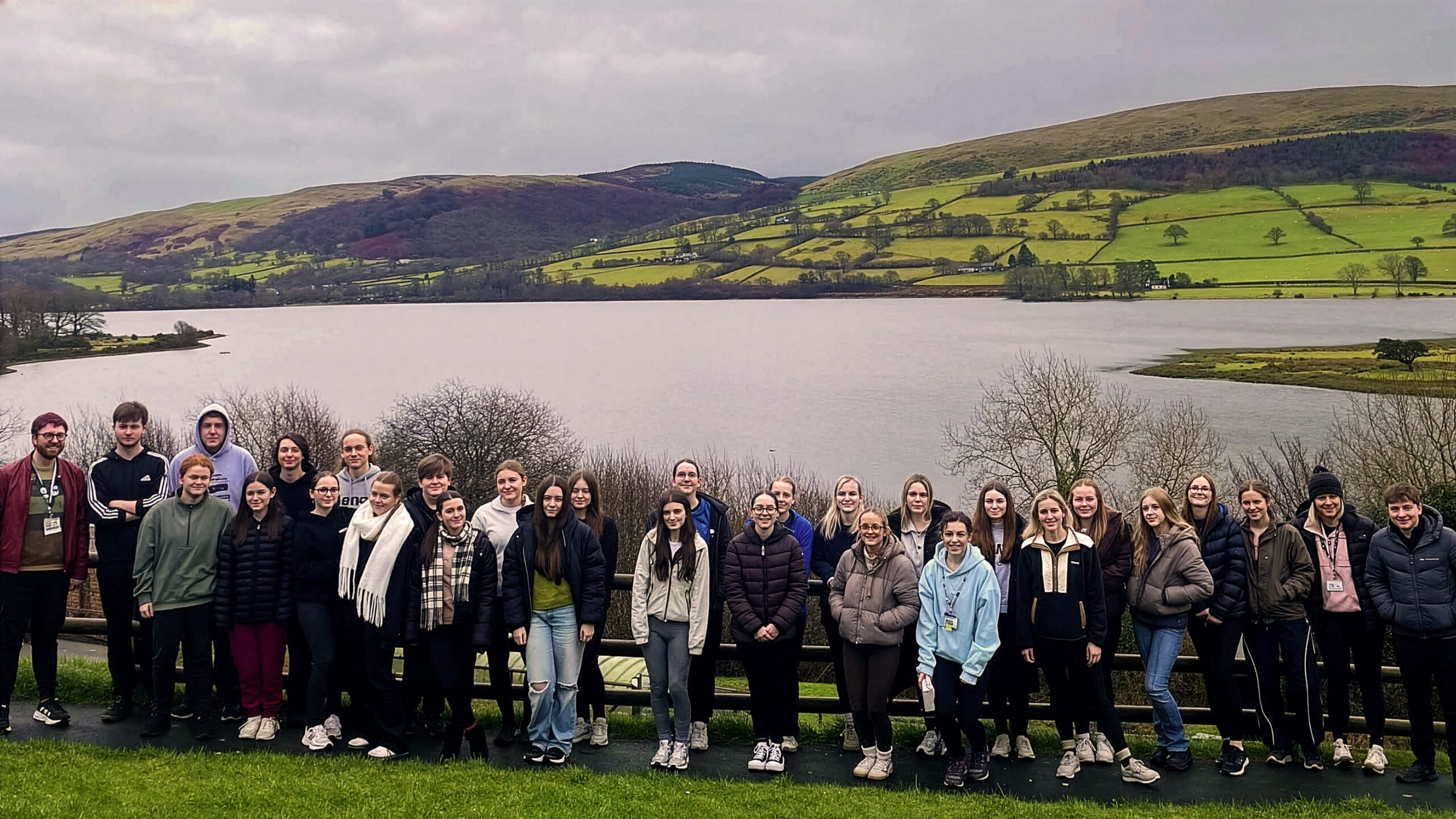 A large group of students and staff standing together by a lakeside viewpoint, with calm water in the foreground and rolling green hills and farmland in the background under an overcast sky.