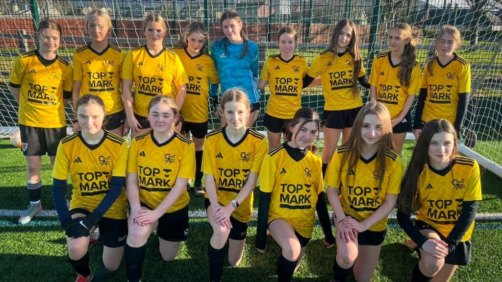 A girls football team group photo on the outdoor pitch, wearing yellow and black kit, circa January 2026