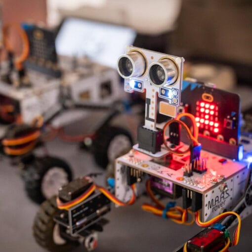 Two small wheeled robots with visible wiring, sensors and LED lights displayed on a table, circa February 2026