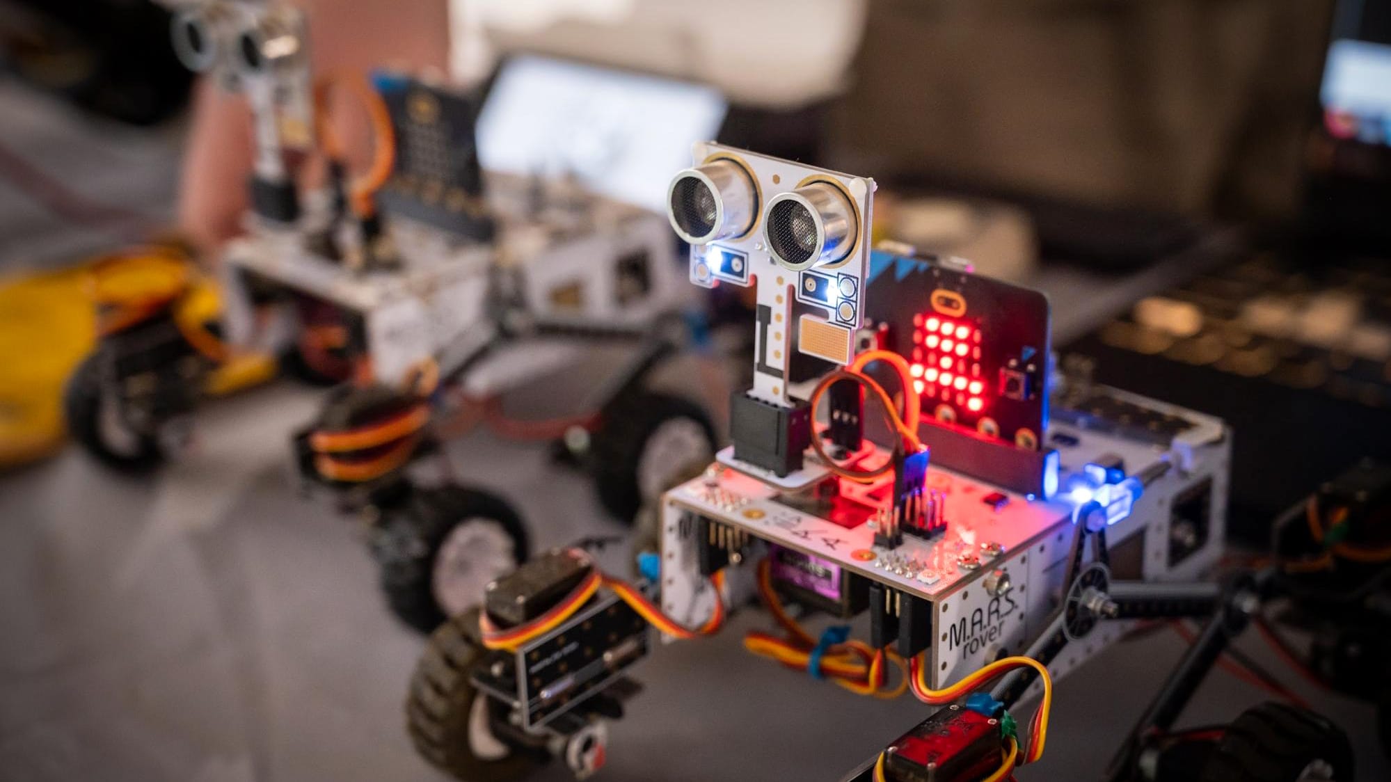 Two small wheeled robots with visible wiring, sensors and LED lights displayed on a table, circa February 2026