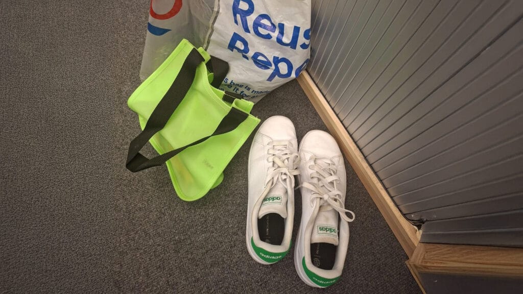 A reusable shopping bag and a bright green bag on the floor next to a pair of white trainers, circa February 2026