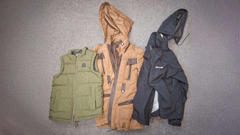 Three coats laid out on a grey carpet: a green padded gilet, a tan hooded coat and a black jacket, circa February 2026