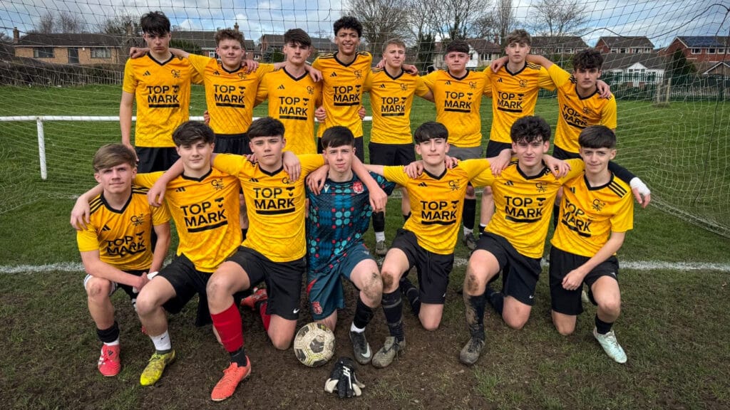 A school football team photo with players in yellow kits posing in front of a goal on a muddy pitch, circa February 2026