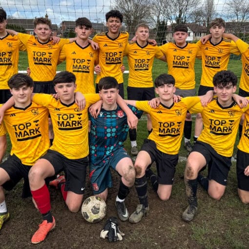 A school football team photo with players in yellow kits posing in front of a goal on a muddy pitch, circa February 2026