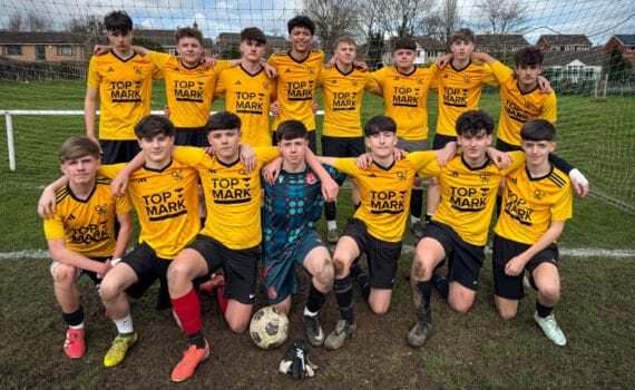 A school football team photo with players in yellow kits posing in front of a goal on a muddy pitch, circa February 2026