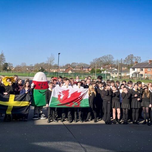 Group of students outdoors holding Welsh and St David's flags alongside a large costume prop during a school event, circa March, 2026