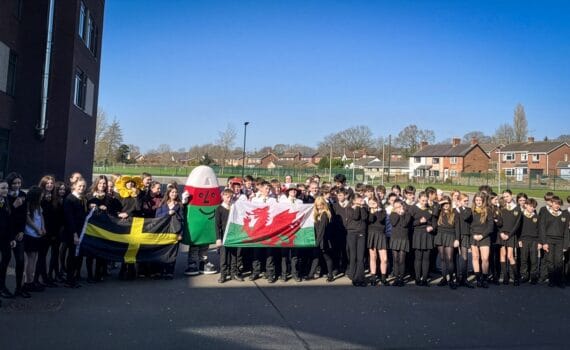 Group of students outdoors holding Welsh and St David's flags alongside a large costume prop during a school event, circa March, 2026