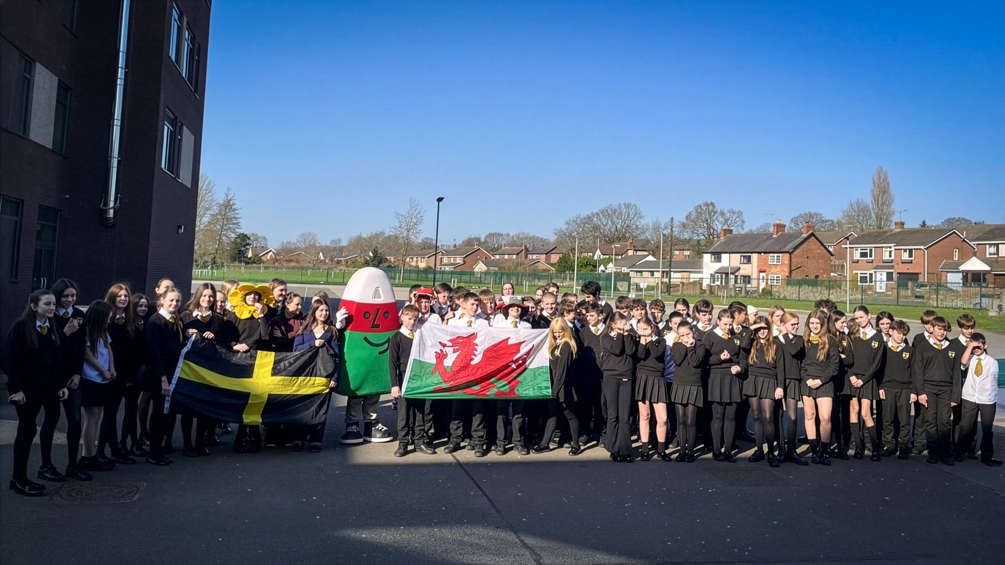 Group of students outdoors holding Welsh and St David's flags alongside a large costume prop during a school event, circa March, 2026