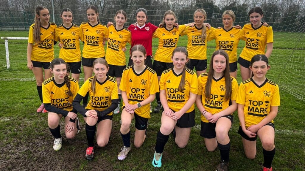 Girls' football team group photo in yellow Castell Alun kits standing and kneeling in front of the goal, circa March, 2026