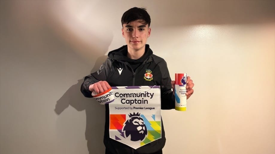 A student stands indoors holding a Premier League Community Captain pennant and award canister while wearing a Wrexham-branded jacket, circa March, 2026