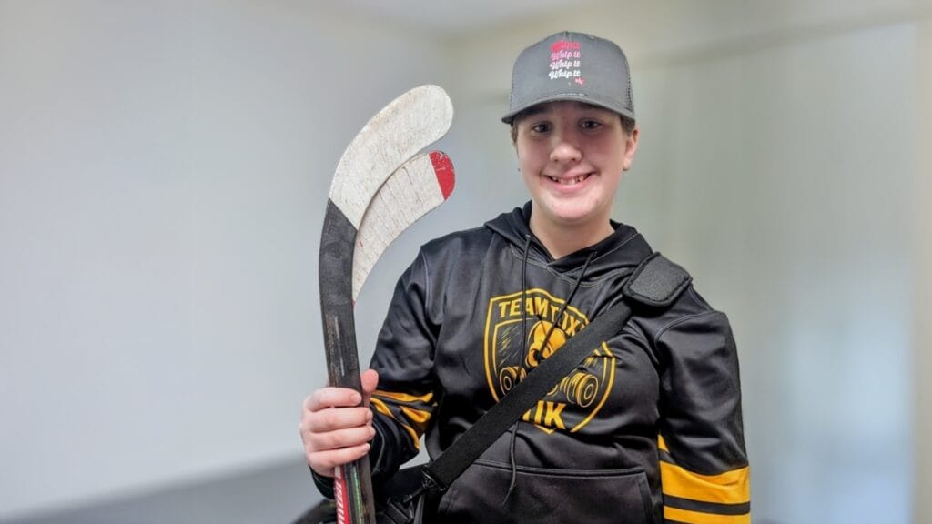 A smiling student in sportswear and a cap holds a hockey stick while posing indoors, circa March, 2026