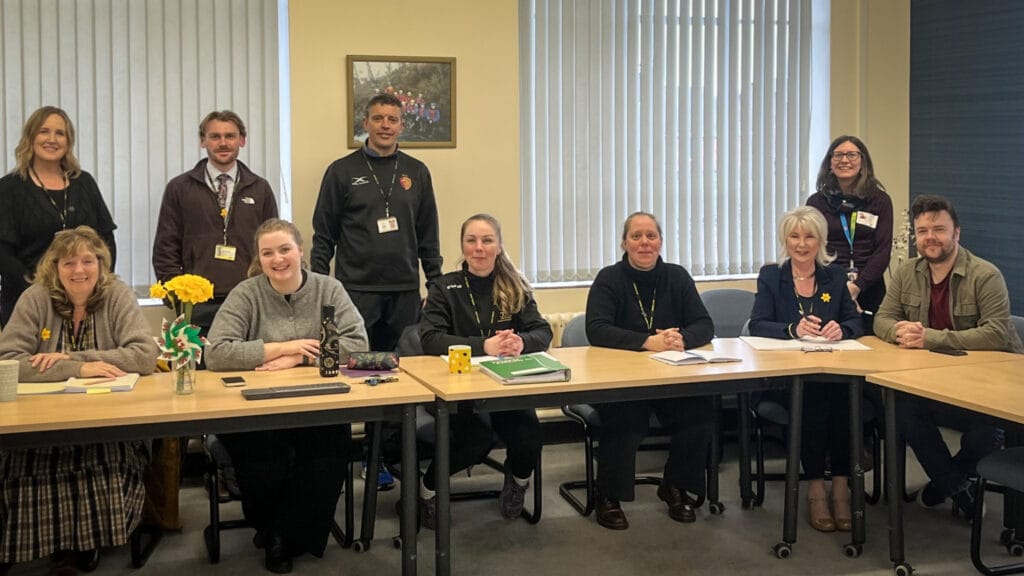 School staff gathered around a meeting table for a group photograph, with daffodils displayed at the front, circa March, 2026