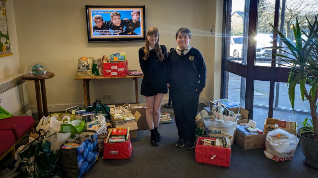 Two pupils standing beside donations of books and learning materials in a school foyer, circa March, 2026