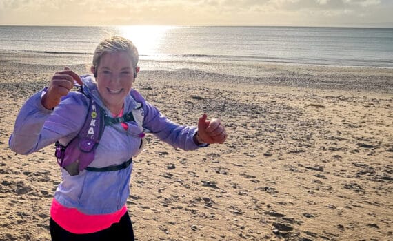 A smiling woman in running gear gives a thumbs-up while jogging along a sandy beach in bright sunshine.