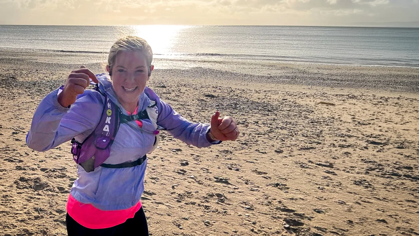 A smiling woman in running gear gives a thumbs-up while jogging along a sandy beach in bright sunshine.