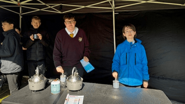 Four students stand behind a table under a gazebo, serving hot drinks from small burners and pouring into cups during an outdoor school event