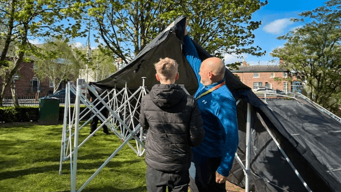 A staff member and a student lift and stretch the frame of a large gazebo while setting it up on a grassy school field with buildings in the background