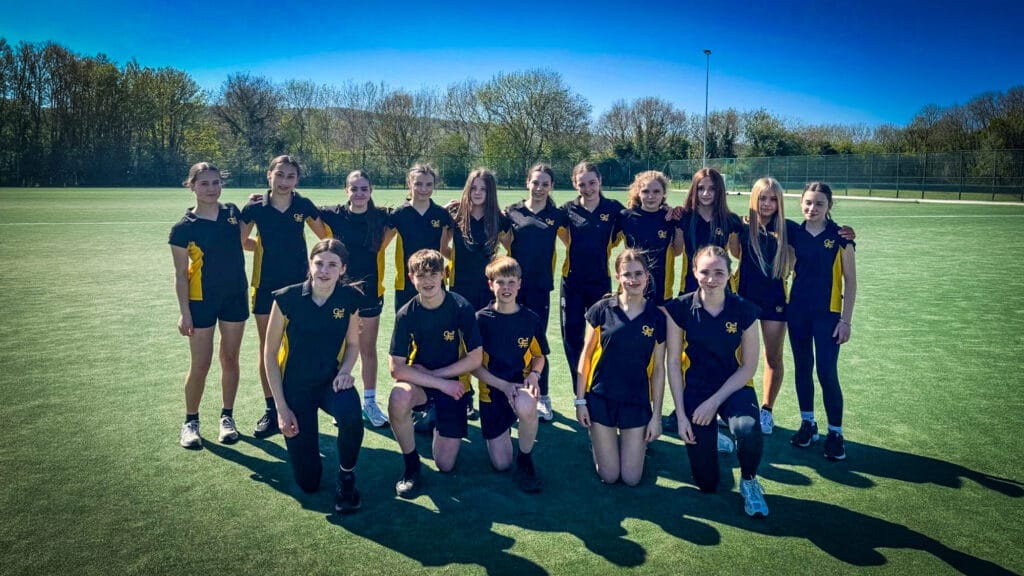 A group of students wearing navy and yellow sports kit pose together on an outdoor artificial pitch, arranged in two rows with some standing and others kneeling, with trees, fencing and a clear blue sky behind them